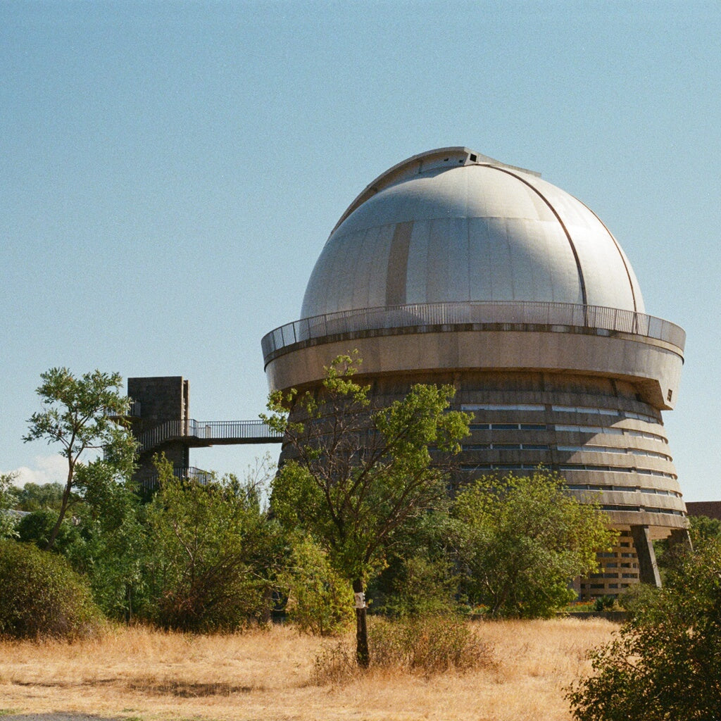 Dome of an observatory surrounded by trees and dry grass under a clear blue sky.